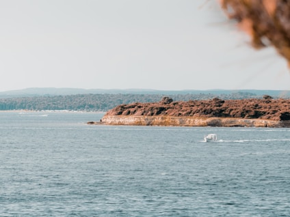 A serene coastal scene featuring a small boat gliding across calm blue waters. The coastline is lush with greenery, and the landscape extends into the distance. The sky is clear, suggesting a peaceful day.