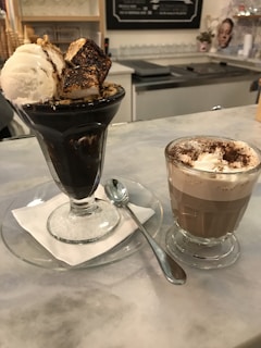 A dessert scene on a countertop featuring a glass filled with dark ice cream topped with toasted marshmallows and a scoop of vanilla ice cream on the side. Adjacent to it is a cup of hot chocolate with whipped cream, sprinkled with cocoa powder. A spoon is placed on the saucer beside the glass.
