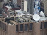 An urban rooftop scene featuring multiple satellite dishes and HVAC units. The view includes a variety of buildings with brick facades, windows, and a prominent mural painted in blue tones on a tall building. Metal railings and industrial equipment are visible on the rooftop.