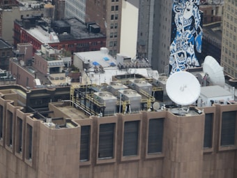 An urban rooftop scene featuring multiple satellite dishes and HVAC units. The view includes a variety of buildings with brick facades, windows, and a prominent mural painted in blue tones on a tall building. Metal railings and industrial equipment are visible on the rooftop.