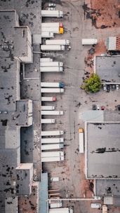 An aerial view of an industrial area with multiple large trucks parked in loading bays next to a large warehouse. The ground appears worn with patches, and there is a small section of greenery. The area contains various vehicles, including a few smaller cars and numerous trailers attached to the trucks.