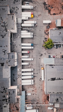 An aerial view of an industrial area with multiple large trucks parked in loading bays next to a large warehouse. The ground appears worn with patches, and there is a small section of greenery. The area contains various vehicles, including a few smaller cars and numerous trailers attached to the trucks.