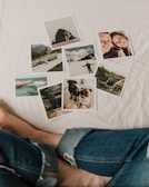 A collection of photographs scattered on a white surface, including landscapes, a couple, and two dogs. The images feature mountains, a coastal scene, a young man in a snowy setting, and close-ups of the dogs. A person's legs in jeans are visible near the photos.