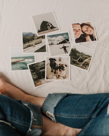 A collection of photographs scattered on a white surface, including landscapes, a couple, and two dogs. The images feature mountains, a coastal scene, a young man in a snowy setting, and close-ups of the dogs. A person's legs in jeans are visible near the photos.
