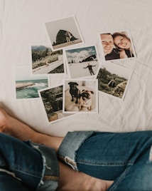 A collection of photographs scattered on a white surface, including landscapes, a couple, and two dogs. The images feature mountains, a coastal scene, a young man in a snowy setting, and close-ups of the dogs. A person's legs in jeans are visible near the photos.