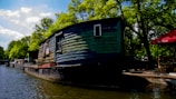 A rustic, wooden houseboat is moored along a canal, surrounded by lush green trees under a partly cloudy sky. The water is calm and reflects the surrounding scenery. Nearby, a red umbrella with cafe seating is visible, suggesting a quaint outdoor dining area.