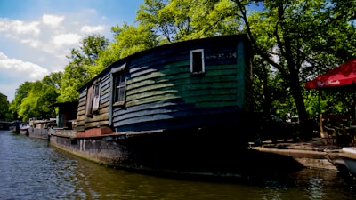 A rustic, wooden houseboat is moored along a canal, surrounded by lush green trees under a partly cloudy sky. The water is calm and reflects the surrounding scenery. Nearby, a red umbrella with cafe seating is visible, suggesting a quaint outdoor dining area.
