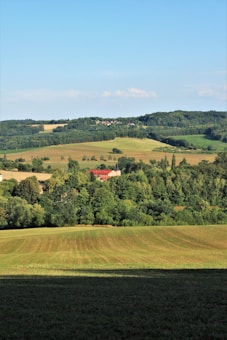 A scenic landscape featuring rolling fields with various shades of green and brown. A cluster of trees surrounds a pair of buildings with red roofs nestled in the middle distance. The background showcases a line of hills covered with dense forests under a clear blue sky.
