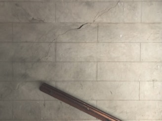 A cracked, pale, tiled floor with a prominent diagonal crack running across the surface. A dark brown wooden bannister is positioned at the bottom left corner, casting a shadow on the floor.