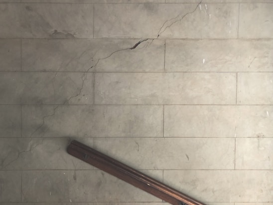 A cracked, pale, tiled floor with a prominent diagonal crack running across the surface. A dark brown wooden bannister is positioned at the bottom left corner, casting a shadow on the floor.