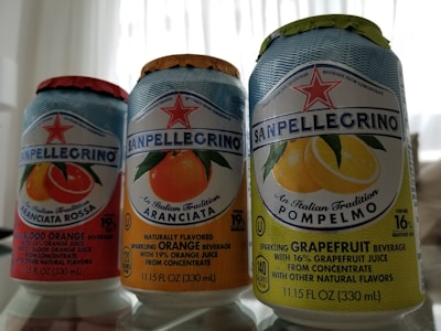 Three cans of Sanpellegrino sparkling beverages are placed side by side, each labeled with different fruit flavors: blood orange, orange, and grapefruit. The design features bright, colorful images of the corresponding fruits with a star logo above the brand name. The cans have details about the juice content and other natural flavors.