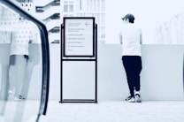 A person stands casually leaning against a low wall, facing away, wearing a white shirt, dark pants, and a cap. Adjacent to the person is a directory sign listing various businesses like a wine bar, hair salon, gallery, and market. The background features modern urban architecture with geometric patterns.