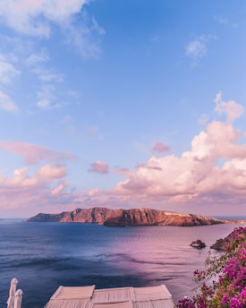 A serene coastal landscape with a view of a rocky island under a vibrant pink and blue sky. Calm waters reflect the sky's colors, and pink bougainvillea flowers decorate the lower right corner. White patio umbrellas are partially visible in the foreground, adding a touch of leisure to the scene.