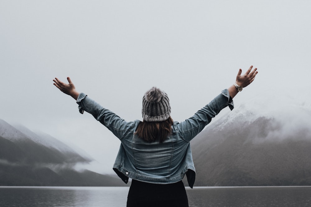 woman lifting both hands while facing body of water and mountain