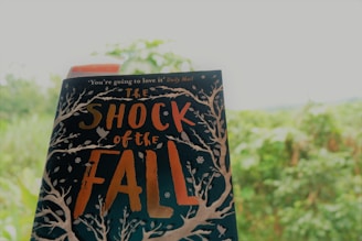 A book titled 'The Shock of the Fall' held up against a blurred background of lush greenery. The book cover features intricate designs of white trees and branches with an orange and white text. A red bookmark is visible at the top of the book.