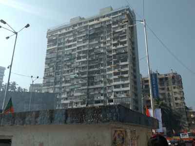 A tall residential building with numerous balconies and windows. Below are street lights, flags, and other buildings, including palm trees in the background. A blue sign reads 'Larsen & Toubro,' adjacent to the main structure.