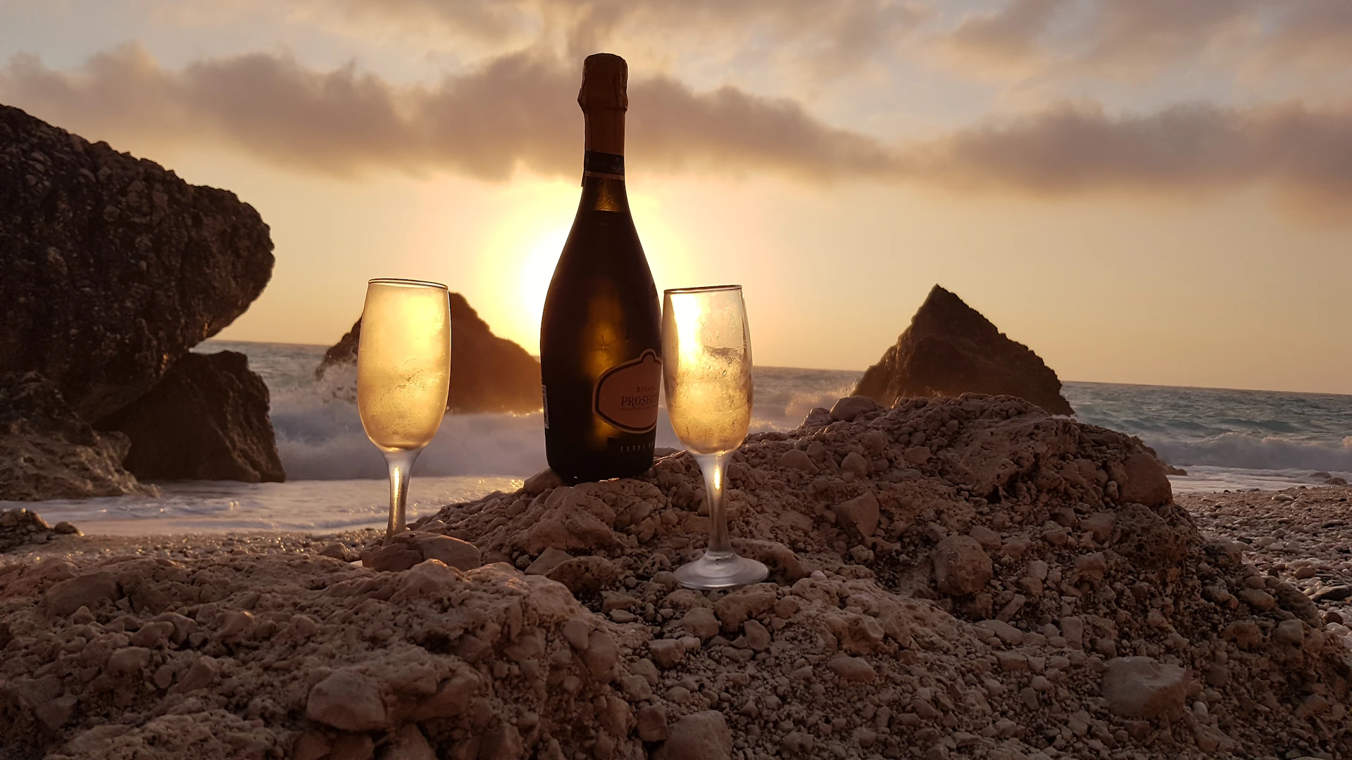 Close-up of friends laughing and clinking their glasses of champagne on the sandy shore as the sun dips below the horizon.
