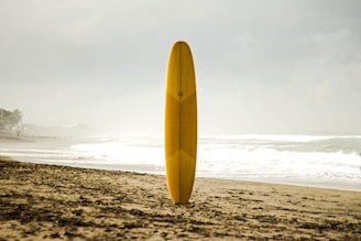 A yellow surfboard stands upright on a sandy beach close to the shoreline. The ocean waves appear active under a cloudy sky, and the shoreline extends into the distance with subtle silhouettes of people and buildings visible.