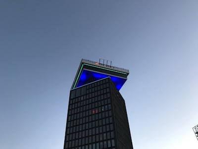 A modern skyscraper with an unusual architectural feature at the top, featuring a large overhanging platform illuminated with blue and green lights against a clear sky.