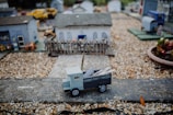 A detailed miniature scene depicting a small rural setting with several scaled-down buildings. At the forefront is a model truck labeled 'Border Farm Timber', carrying small logs. Gravel paths and tiny structures, including a fenced yard, add depth to the scene.