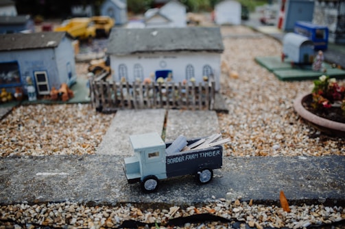 A detailed miniature scene depicting a small rural setting with several scaled-down buildings. At the forefront is a model truck labeled 'Border Farm Timber', carrying small logs. Gravel paths and tiny structures, including a fenced yard, add depth to the scene.