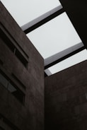 An architectural view of a modern building featuring geometric shapes, with a partially open roof displaying the sky. The structure is composed of dark stone or concrete panels, giving it an industrial appearance.