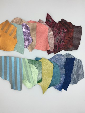 A collection of colorfully patterned fabric or leather swatches arranged on a flat, white surface. The swatches vary in color, including shades of orange, pink, teal, gold, red, purple, and blue, with textures ranging from smooth to snakeskin-like patterns. Some pieces feature metallic sheens or stripes.