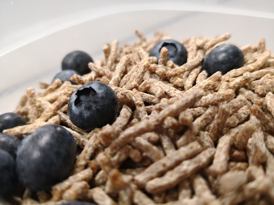 A close-up view of a bowl filled with breakfast cereal sticks and blueberries. The blueberries are scattered among the crunchy, fibrous cereal pieces, adding a splash of dark blue color and a contrast in texture.