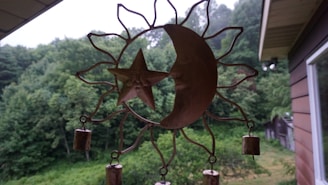 A metal wind chime with a sun and moon design hangs against a backdrop of lush, green trees. The chime consists of a crescent moon and a star with a face inside a sun-shaped frame. Small cylindrical bells are attached to the bottom.