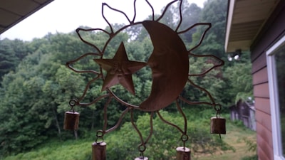 A metal wind chime with a sun and moon design hangs against a backdrop of lush, green trees. The chime consists of a crescent moon and a star with a face inside a sun-shaped frame. Small cylindrical bells are attached to the bottom.