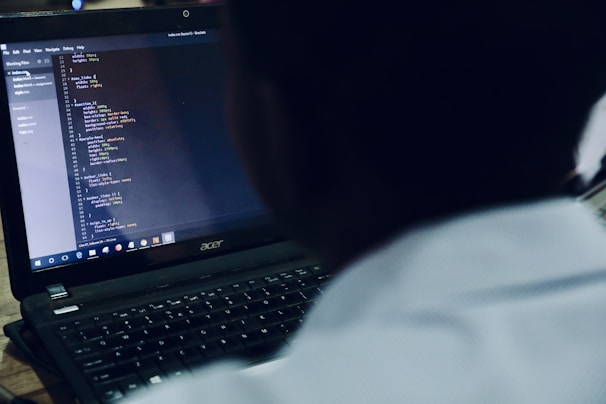 A person is focused on programming code displayed on a laptop screen. The code is organized in a text editor, and the viewer is looking over the shoulder of the person at the screen.