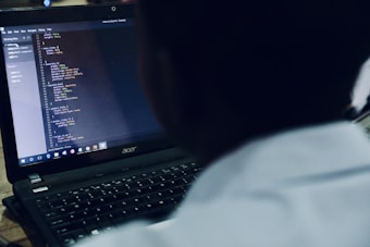 A person is focused on programming code displayed on a laptop screen. The code is organized in a text editor, and the viewer is looking over the shoulder of the person at the screen.