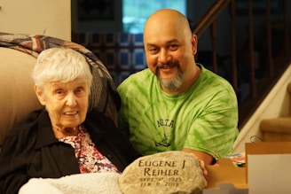 An elderly woman and a man are seated together, smiling at the camera. The woman has short white hair and is wearing a floral top with a dark jacket. The man is wearing a green T-shirt and is holding a stone with engraved text that reads 'Eugene J. Reiher, Sept. 17, 1925 - Feb. 9, 2013.' The setting appears to be indoors, with a staircase in the background.