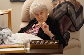 An elderly woman with short white hair is seated comfortably in a beige chair, holding a piece of chocolate from an open box of assorted chocolates. She is wearing a floral top under a dark cardigan. Beside her, a wooden side table holds a glass of water and a tissue box. A patterned blanket drapes over the back of the chair, contributing to a cozy atmosphere.