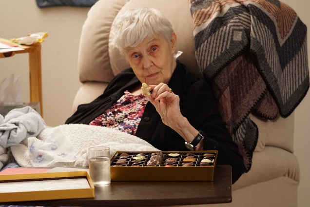 An elderly woman with short white hair is seated comfortably in a beige chair, holding a piece of chocolate from an open box of assorted chocolates. She is wearing a floral top under a dark cardigan. Beside her, a wooden side table holds a glass of water and a tissue box. A patterned blanket drapes over the back of the chair, contributing to a cozy atmosphere.