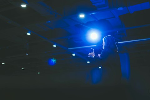 A person appears to be delivering a speech or presentation under bright stage lights in a dimly lit auditorium. The scene is dominated by a strong blue light casting a dramatic effect over the speaker and surroundings.
