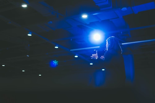 A person appears to be delivering a speech or presentation under bright stage lights in a dimly lit auditorium. The scene is dominated by a strong blue light casting a dramatic effect over the speaker and surroundings.