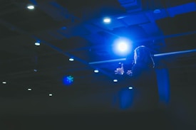 A person appears to be delivering a speech or presentation under bright stage lights in a dimly lit auditorium. The scene is dominated by a strong blue light casting a dramatic effect over the speaker and surroundings.