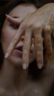 A close-up of a hand with long, natural nails, covered in a glossy, sticky substance. The focus is on the fingers, which are spread apart. In the background, part of a face is visible, slightly out of focus.