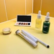 A selection of beauty products is displayed on a yellow countertop. There is an eyeshadow palette with neutral shades, a round compact with a golden lid, two dropper bottles containing serums, a makeup brush with a gold handle, and two white pen-shaped makeup products.