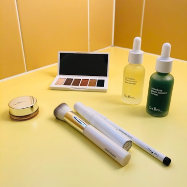A selection of beauty products is displayed on a yellow countertop. There is an eyeshadow palette with neutral shades, a round compact with a golden lid, two dropper bottles containing serums, a makeup brush with a gold handle, and two white pen-shaped makeup products.