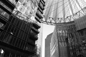 A complex architectural scene featuring tall, modern buildings with extensive use of glass and steel. The structures frame a geometric roof made of steel beams and translucent panels, casting intricate shadows and reflections. The perspective emphasizes height and modernity.