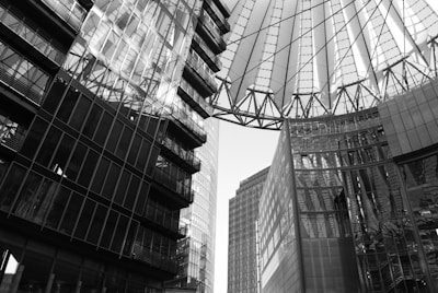 A complex architectural scene featuring tall, modern buildings with extensive use of glass and steel. The structures frame a geometric roof made of steel beams and translucent panels, casting intricate shadows and reflections. The perspective emphasizes height and modernity.