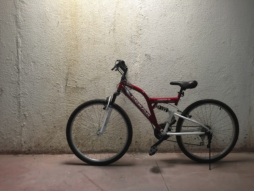 A red and white mountain bike with a sturdy frame and black seat is leaned against a rough-textured concrete wall. The lighting is dim with focused illumination highlighting the bike against the nearly featureless, beige wall.