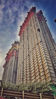 Two towering skyscrapers under construction reach into a partially cloudy sky. The buildings are surrounded by scaffolding, with numerous windows arranged symmetrically across their facades. At the base, lush green trees contrast sharply with the towering structures.