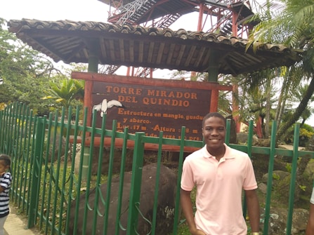 A young man wearing a light pink shirt stands smiling in front of a wooden sign that reads 'TORRE MIRADOR DEL QUINDIO.' The sign is mounted under a small roof structure supported by poles. Green fencing surrounds the area, and lush greenery, including palm trees, is visible in the background. A child in a striped shirt is partially visible to the left.