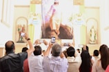 A group of people is gathered in a church or religious setting, holding up smartphones to take photos of a large religious painting. The space is adorned with statues and religious symbols around the painting.