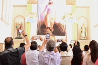 A group of people is gathered in a church or religious setting, holding up smartphones to take photos of a large religious painting. The space is adorned with statues and religious symbols around the painting.