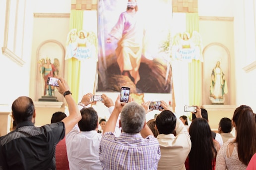 A group of people is gathered in a church or religious setting, holding up smartphones to take photos of a large religious painting. The space is adorned with statues and religious symbols around the painting.