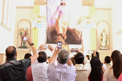 A group of people is gathered in a church or religious setting, holding up smartphones to take photos of a large religious painting. The space is adorned with statues and religious symbols around the painting.